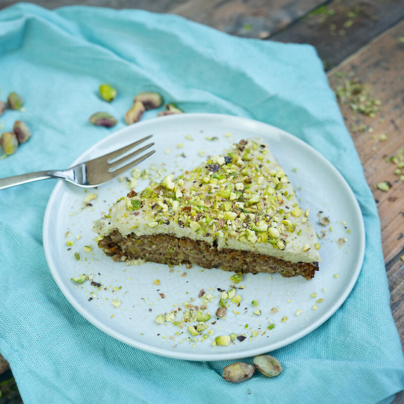 Pistazienkuchen Ein dreieckiges Stück Pistazienkuchen mit heller Cashew-Glasur, bestreut mit grob gehackten Pistazien, liegt auf einem weißen Teller auf einem türkisfarbenen Tuch. Eine Gabel liegt daneben.