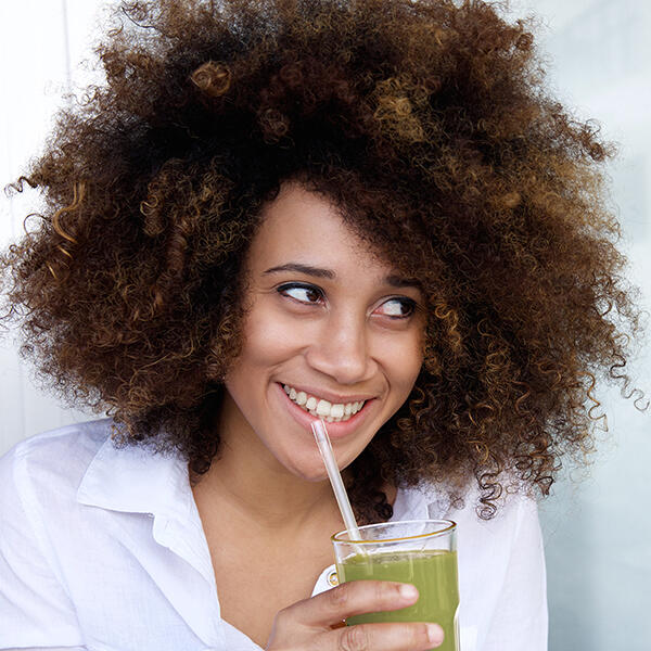 Eine Frau mit lockigem Haar trinkt lächelnd einen grünen Smoothie durch einen Strohhalm.