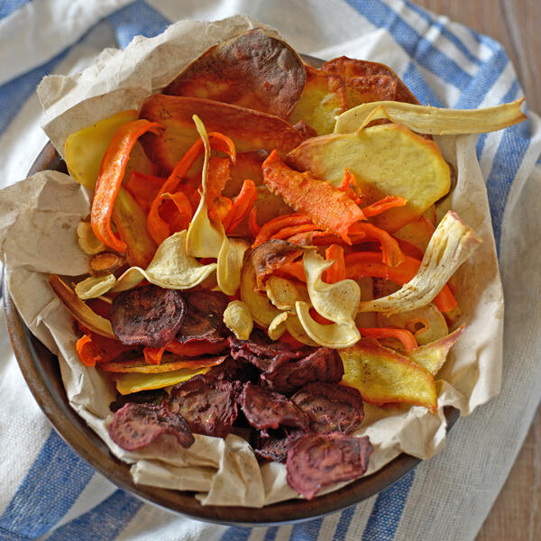 Verschiedene bunte, dünn geschnittene Gemüsescheiben liegen auf Backpapier in einer Schale und wirken getrocknet und knusprig.