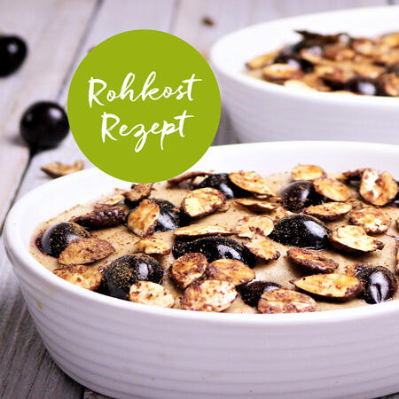 Weiße Schale mit roh veganer Süßkirschen-Creme, bedeckt mit gerösteten Mandel- und Cashewkernscheiben auf Holztisch. Grüner Kreis mit weißer Schrift Rohkost Rezept.