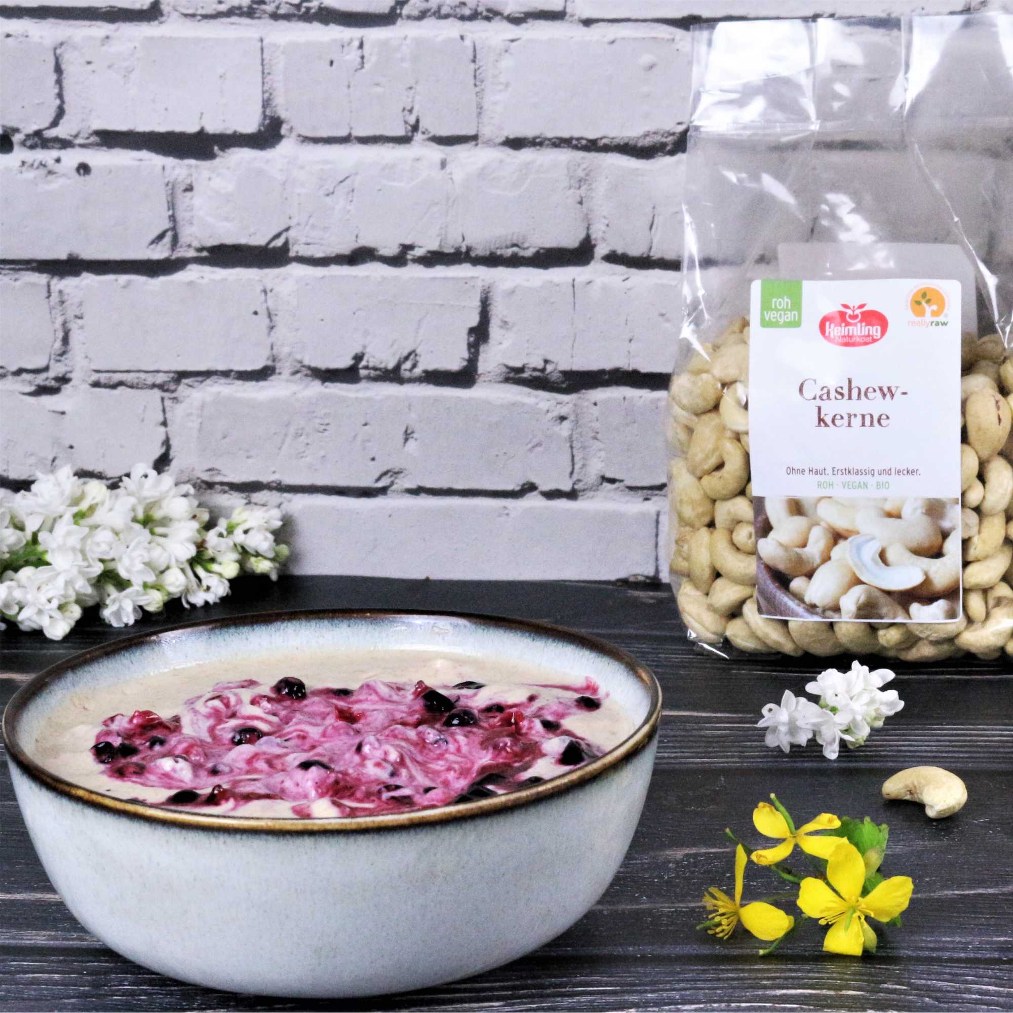 Grießpudding mit Vanille & schwarzen Johannisbeeren, roh-vegan Schüssel mit Grießpudding, verfeinert mit Vanille und schwarzen Johannisbeeren, dahinter eine Packung roher, veganer Cashewkerne von Heimling Naturkost. Weiße und gelbe Blüten liegen dekorativ auf dem dunklen Holz.