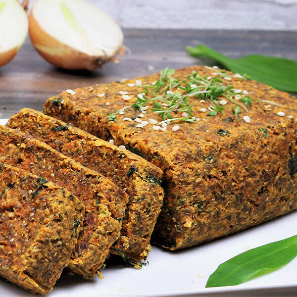 Bärlauchbrot, roh-vegan Das vegane Bärlauchbrot liegt auf einem weißen Teller und ist teilweise in Scheiben geschnitten. Das Brot ist grobkörnig und mit frischem Bärlauch und Sesam bestreut. Im Hintergrund sind halbierte Zwiebeln zu sehen.