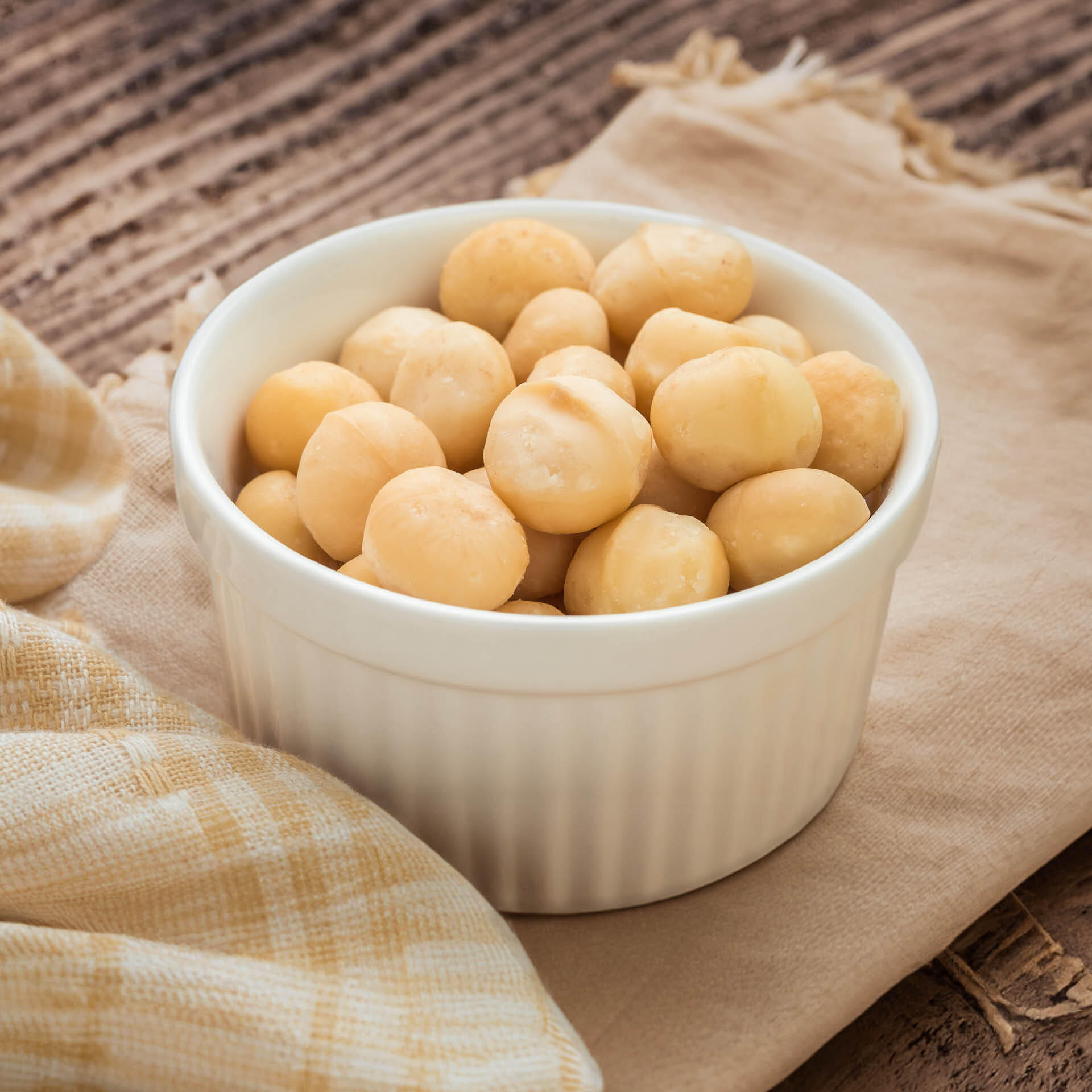 Macadamia, ganze Nüsse, bio 2.5 kg Eine weiße Schale ist mit hellgelben Macadamianüssen gefüllt. Sie steht auf einem beige-hellbraunen Tuch und einem Holztisch im Hintergrund.