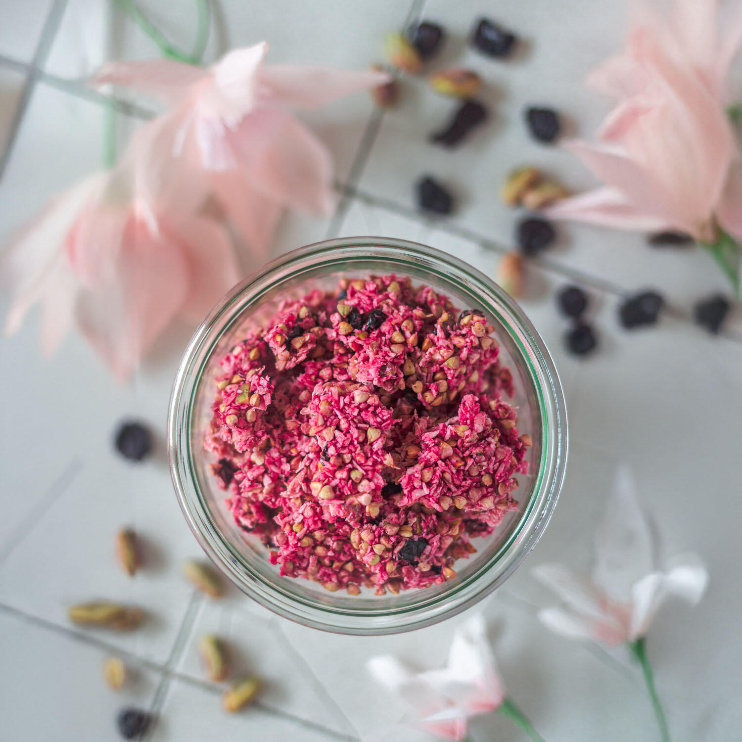 Pink Rawnola Ein Glas mit pinkem Rawnola-Granola aus gekeimtem Buchweizen, Pistazien, Kokos, Kirschen und Vanille steht auf hellen Fliesen. Drumherum liegen rosa und weiße Blüten sowie verstreute Nüsse und getrocknete Beeren.