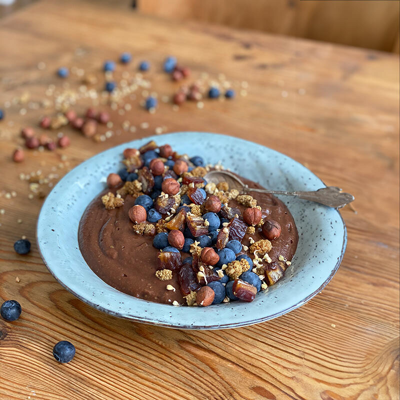 No-tella by Maria Sperling Schokoladen-Bowl mit einer cremigen No-tella-Basis, garniert mit Haselnüssen, Blaubeeren, Datteln und weißen Maulbeeren. Sie steht auf einem Holztisch mit verstreuten Beeren und Nüssen.