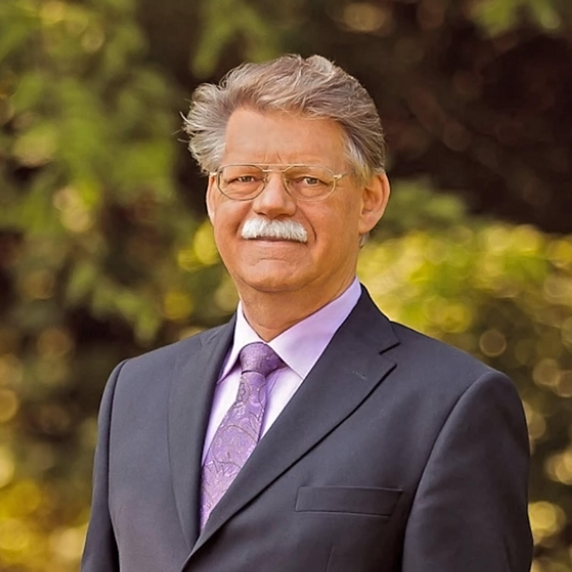 P. Jentschura steht im Anzug mit Fliege vor einem unscharfen, grünen Hintergrund. Er trägt eine Brille und hat graues Haar mit schnurrbartähnlichem Bart.